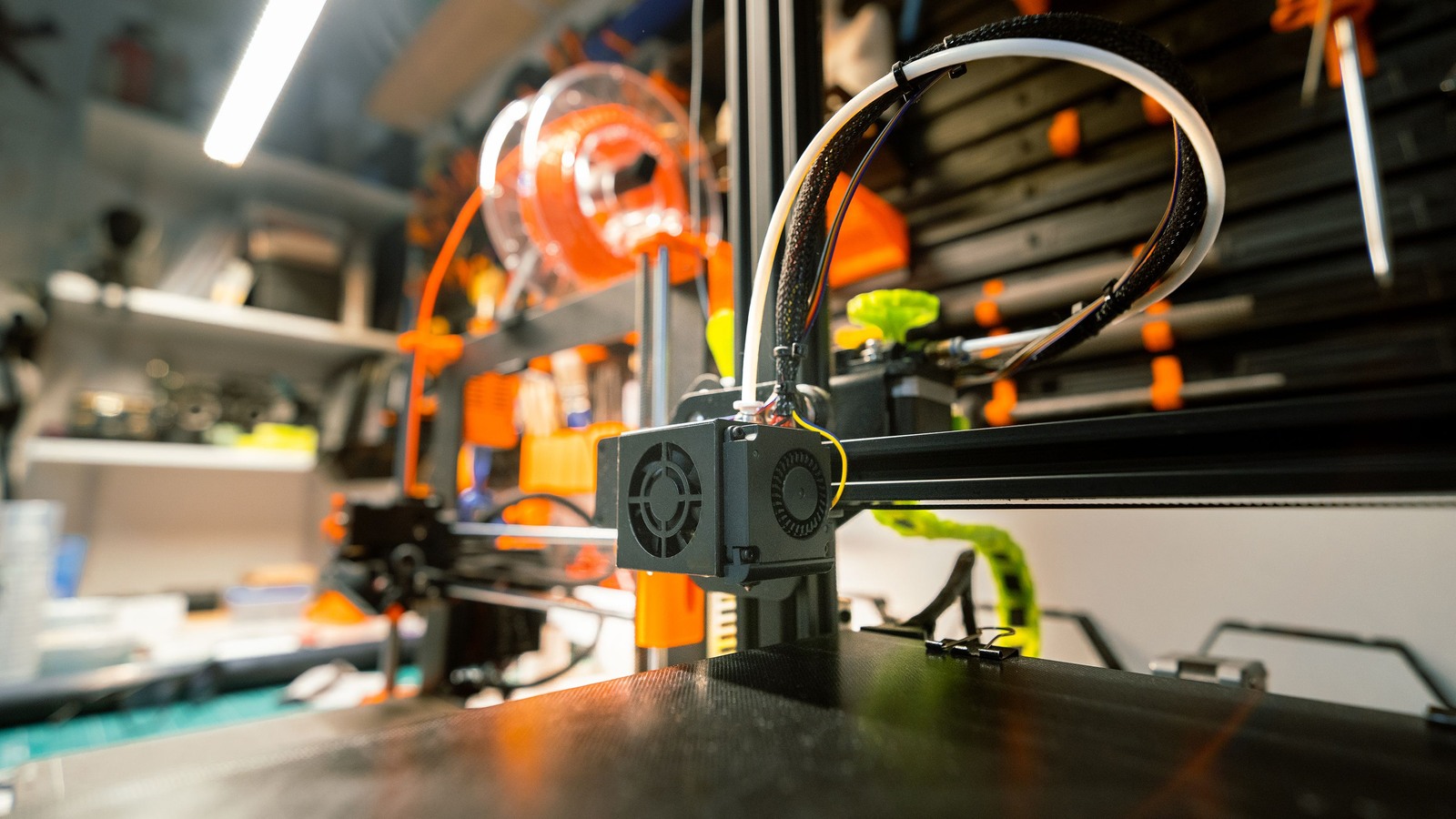 Filament printer toolhead above a build plate, with a spool and workshop tools in the background.