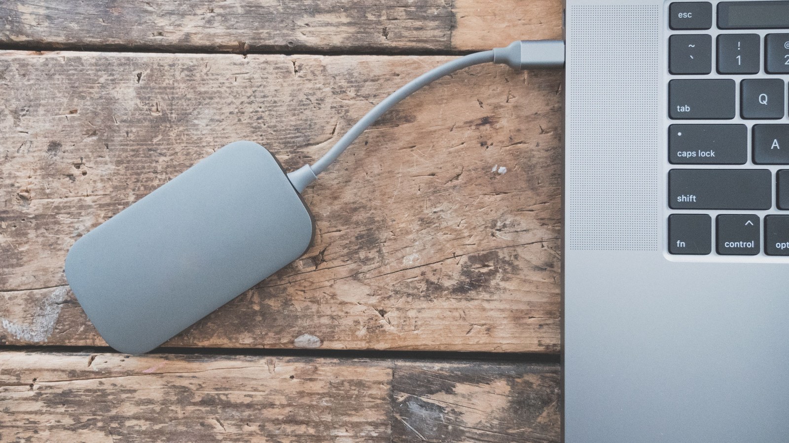 A grey external drive connected by a cable to a laptop on a wooden table.